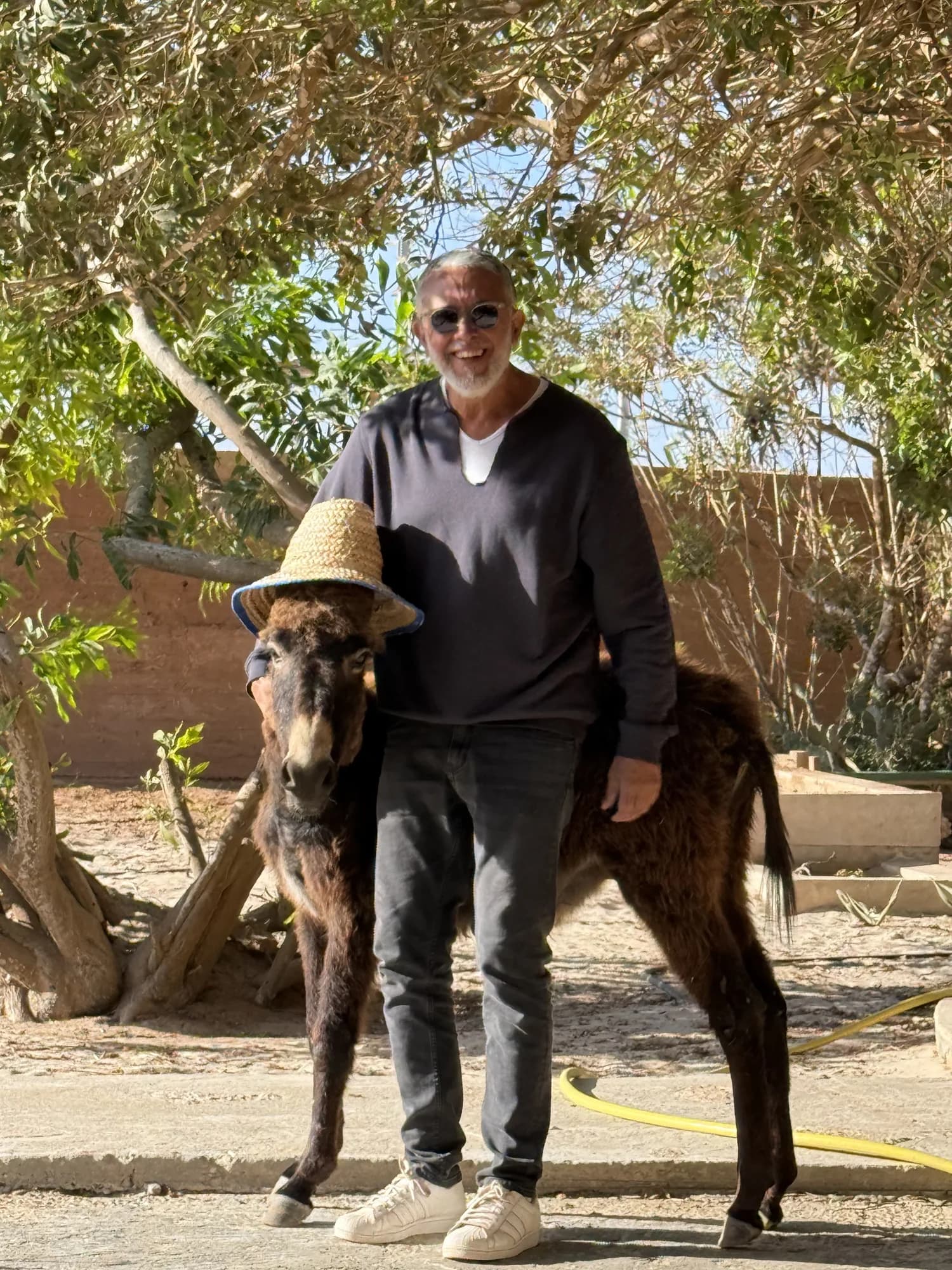 Farid the host, smiling in sunglasses beside his straw-hatted donkey under the garden trees